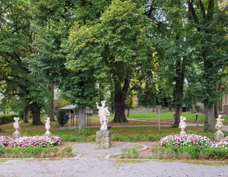 Ein Ausschnitt eines Parks, mit einem gekiesten Weg im Vordergrund , einer großen männlichen Statur und links und rechts jeweils 2 steinerne Engelfiguren