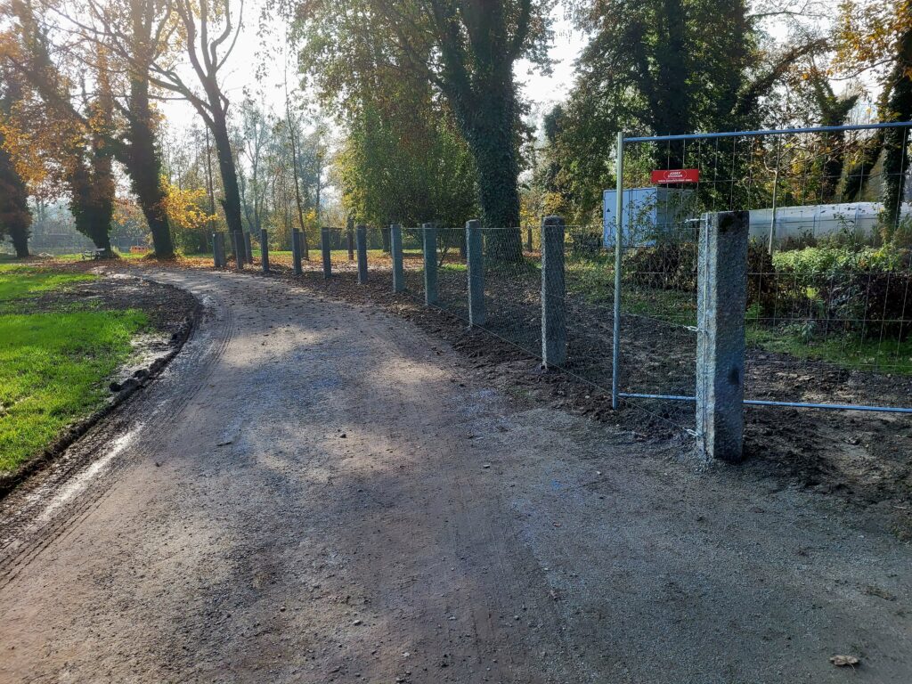 In einem Park mit großen Bäumen im Herbst führt ein Weg. Dieser hat an der rechten Seite einen Zaun aus massiven Steinpfosten und einem Metallgitter.