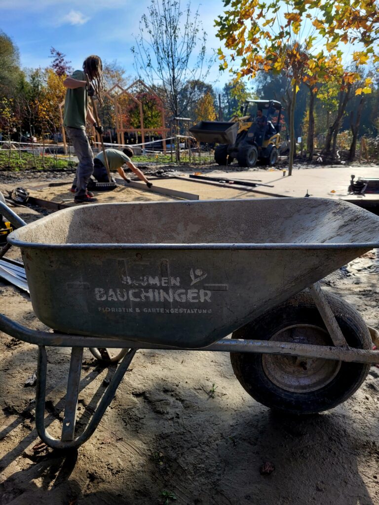 Im Vordergrund ist eine Schubkarre, im Hintergrund arbeiten 2 Männer auf einer Baustelle im Garten.