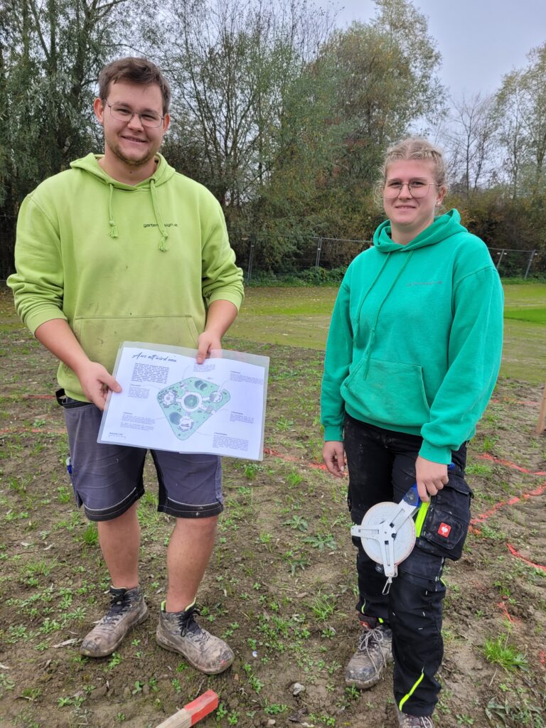 2 junge Landschaftsgärtner in Arbeitskleidung lächeln in die Kamera und befinden sich auf einer Baustelle. Sie halten einen Plan und ein Messrad in der Hand