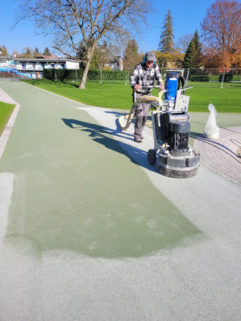 Ein Mann mit Gehörschutz bewegt eine große Schleifmaschine über einen Weg mit zuerst grünen Asphalt und schleift die Oberfläche so ab, dass die Farbe danach hellgrün-grau ist.