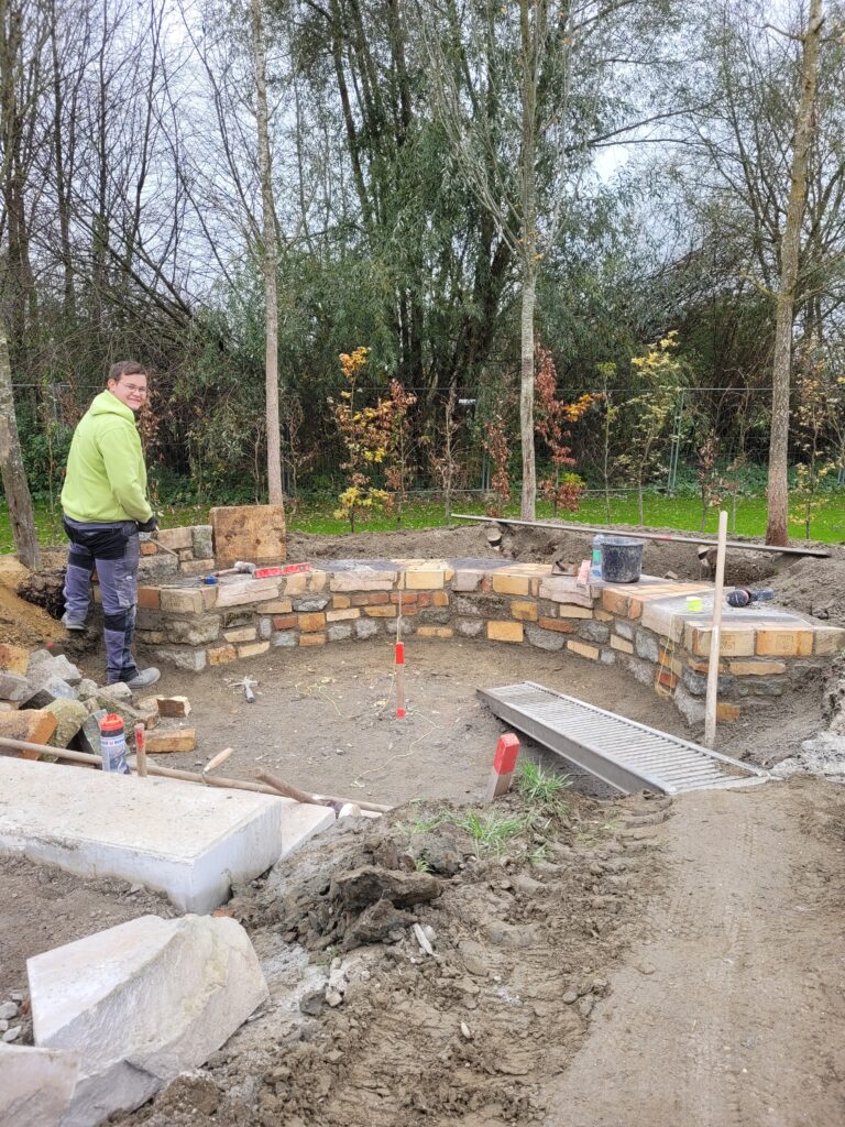 Ein junger Handwerker steht lächelnd vor einer kniehohen Steinmauer, die er aus alten Steinen gebaut hat