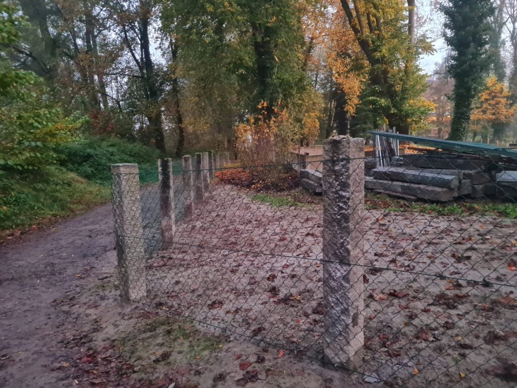 In Großaufnahme ist ein Zaun aus Granitpfosten mit einem Metallgitter in einem Park abgebildet. Die Bäume im Park sind herbstlich gefärbt.