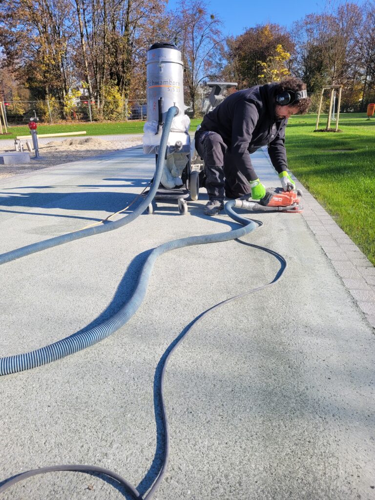 Ein junger Handwerker kniet auf einem asphaltierten Weg, denn er mit einem Handschleifgerät bearbeitet. Er trägt einen Gehörschutz, Handschuhe und Arbeitskleidung.