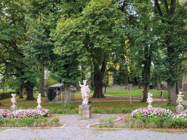 Ein Ausschnitt eines Parks, mit einem gekiesten Weg im Vordergrund , einer großen männlichen Statur und links und rechts jeweils 2 steinerne Engelfiguren