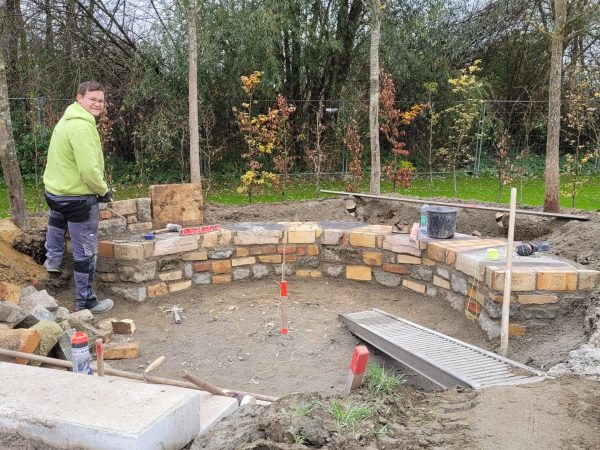 Ein junger Handwerker steht lächelnd vor einer kniehohen Steinmauer, die er aus alten Steinen gebaut hat