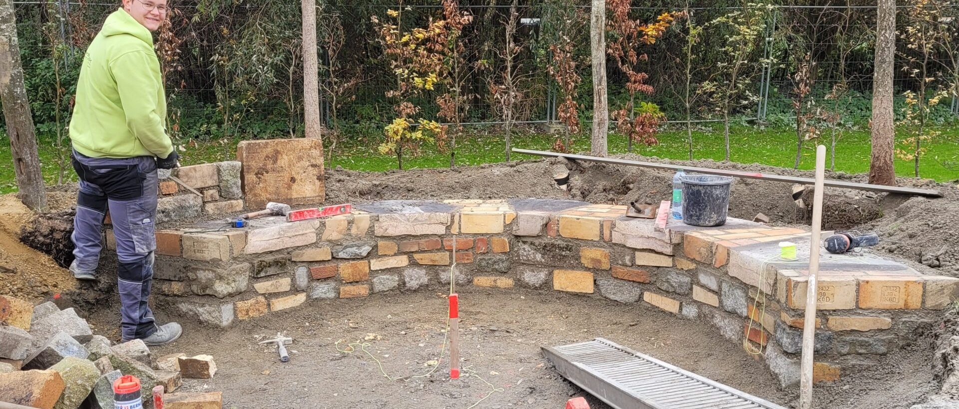 Ein junger Handwerker steht lächelnd vor einer kniehohen Steinmauer, die er aus alten Steinen gebaut hat