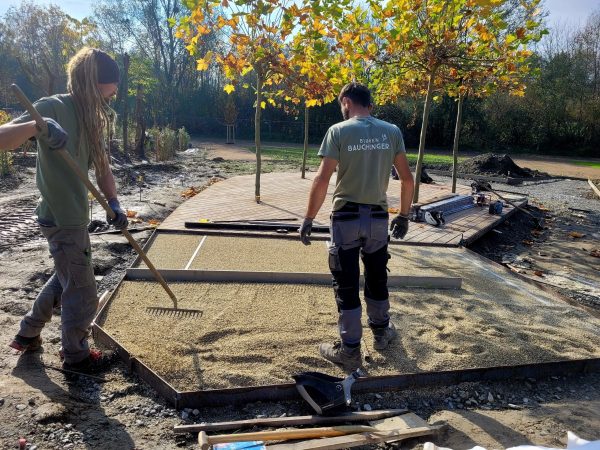 2 Männer arbeiten auf einer Baustelle eines Gartens und bereiten den Boden mit Kies und Rechen vor, damit die Terrassenbretter verlegt werden können.