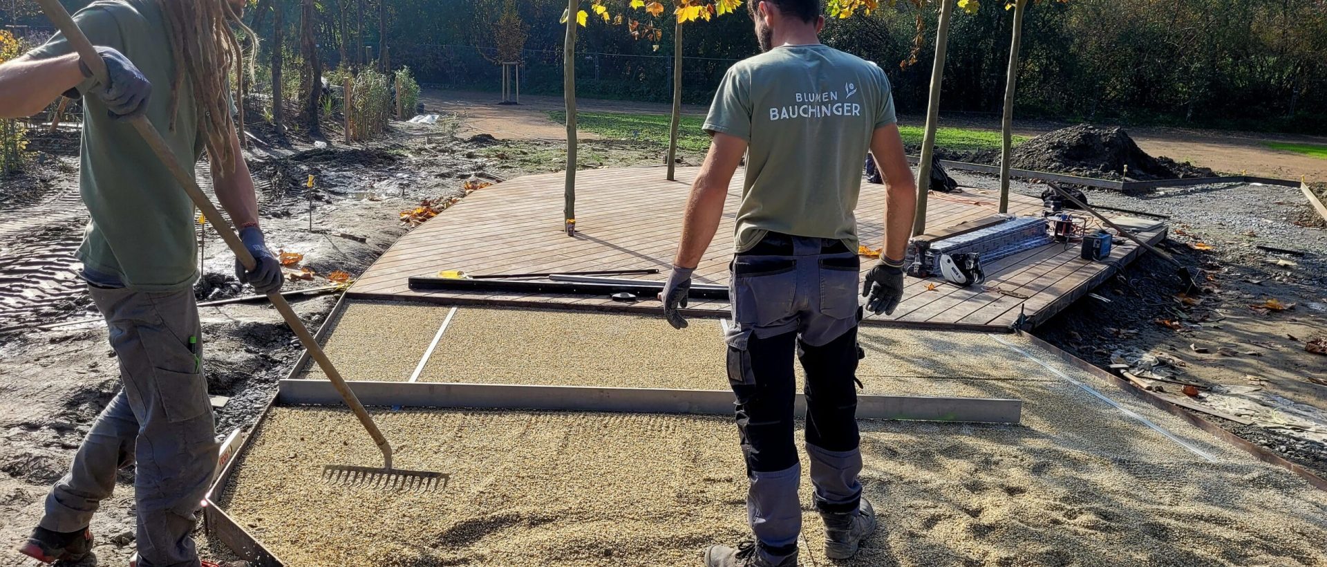 2 Männer arbeiten auf einer Baustelle eines Gartens und bereiten den Boden mit Kies und Rechen vor, damit die Terrassenbretter verlegt werden können.